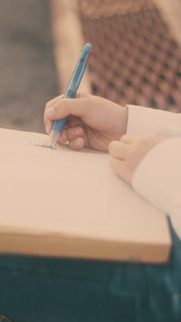 cute schoolboy in glasses draws in paper notebook with pen holding on friend back in sunny street close view