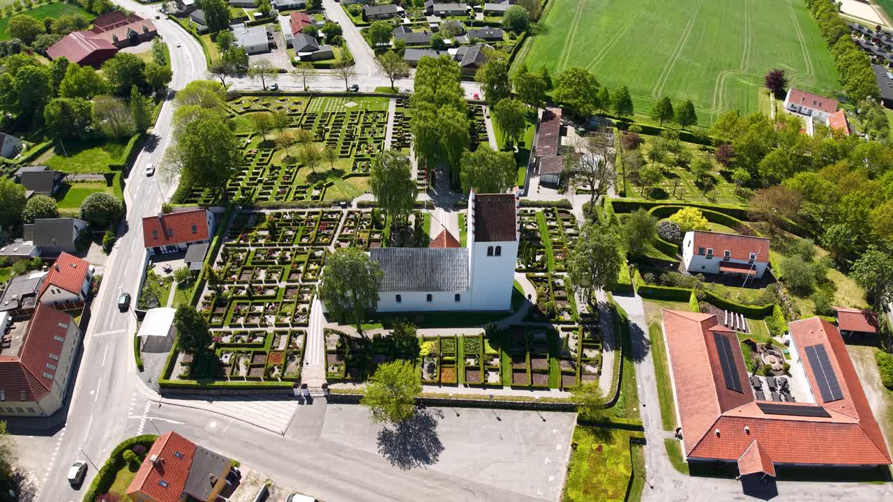 Aerial drone footage of a Danish village church and surrounding cemetery, showing traditional architecture, green gardens, and nearby homes under bright daylight