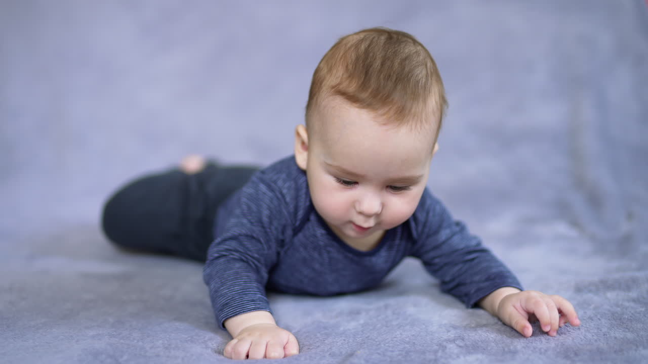 Adorable Caucasian boy in blue clothes lies on belly waving his feet. Nice curious toddler looks up with interest sweetly rising his eyebrows.