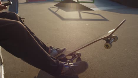 la cámara se enfoca en un joven sentado en un parque de patinaje, mientras su amigo salta en patineta