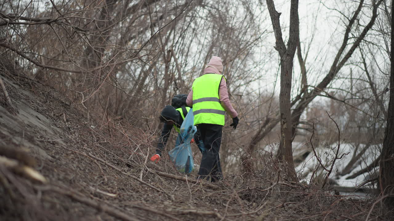 Volunteers in reflective vests and gloves walking along forested riverbank during environmental cleanup, carrying blue plastic bags filled with collected trash to protect nature