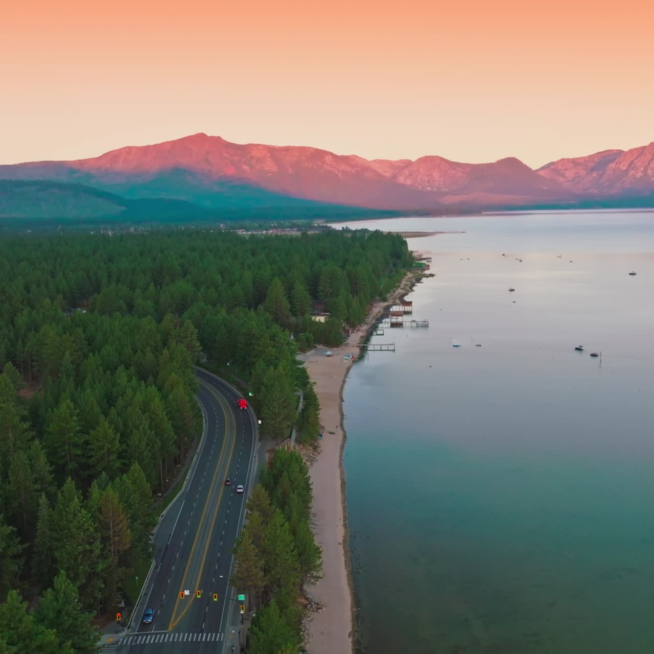 Wooded shore of the Lake Tahoe with wide road through the forest. Pink mountains and pink skies at backdrop. Top view