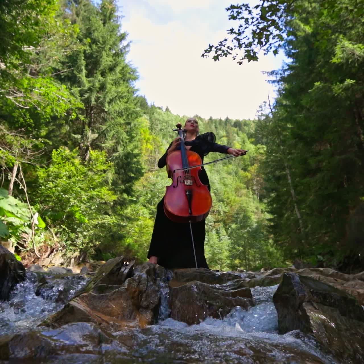 Luxury woman with musical instrument outdoors. Female musician performing music with a cello on amazing nature background with fresh mountain water