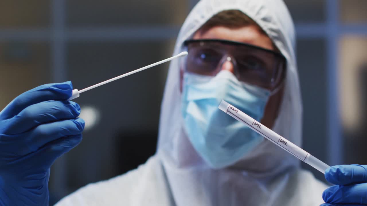 Operatore sanitario caucasico di sesso maschile che indossa indumenti protettivi, mascherina e guanti, mentre ispeziona un tampone di DNA in laboratorio.