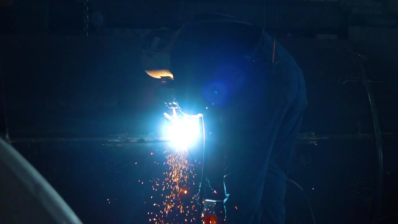Wide slow motion shot of a worker welding on some metal with sparks coming down and a lens flare. 500fps