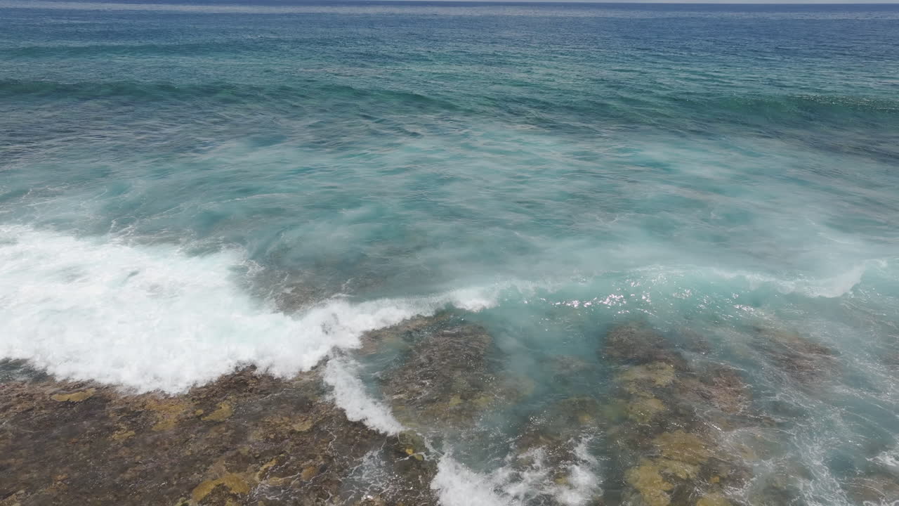 Drone high angle pullback follows crashing waves water over fringing reef with tendrils of whitewash foam spreading, South Pacific, Rarotonga Cook Islands