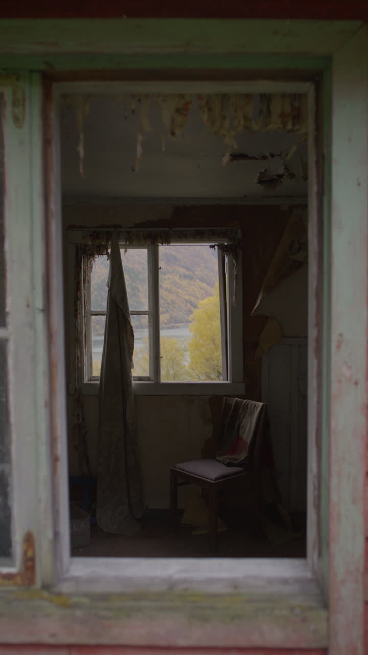 An abandoned house with peeling walls, a tattered curtain, and a chair, overlooking a fjord in Norway. Filmed vertically.