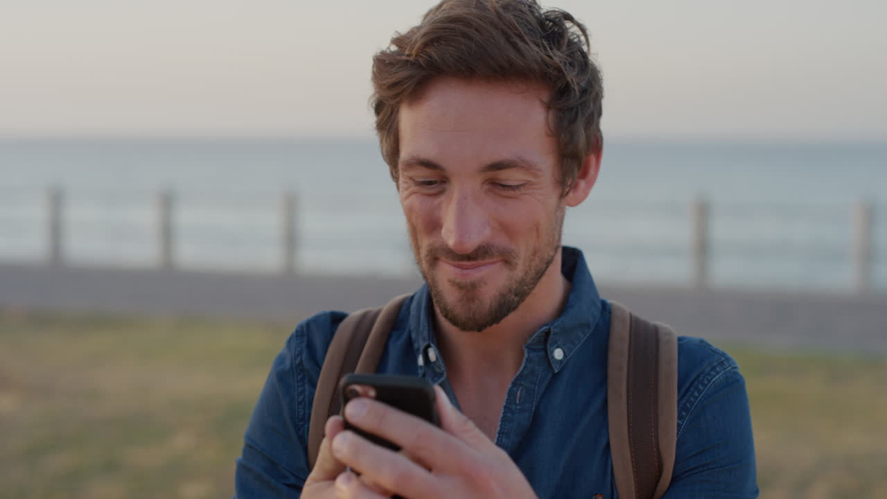 ritratto bel giovane che usa lo smartphone sorridendo godendosi la lettura di messaggi online navigando sull'app del telefono cellulare in slow motion sul mare