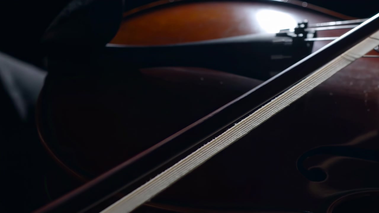 Primo piano di archetto di violino che scivola sulle corde in uno studio in penombra, legno verniciato caldo sotto un riflettore soffuso, lento