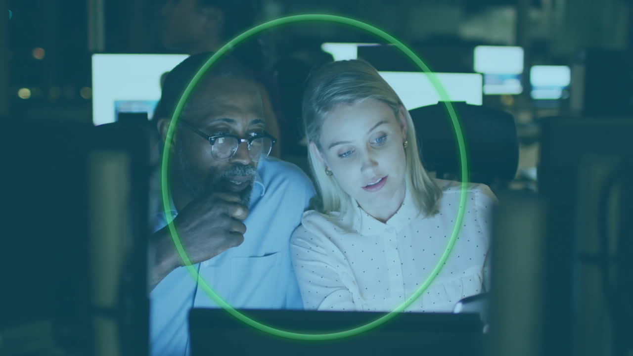 Technology team collaborating on laptop in control room, viewing monitors with green overlay