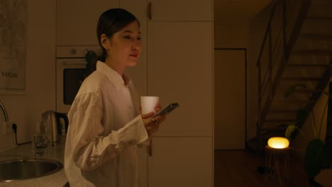 woman drinking and using phone in kitchen at night