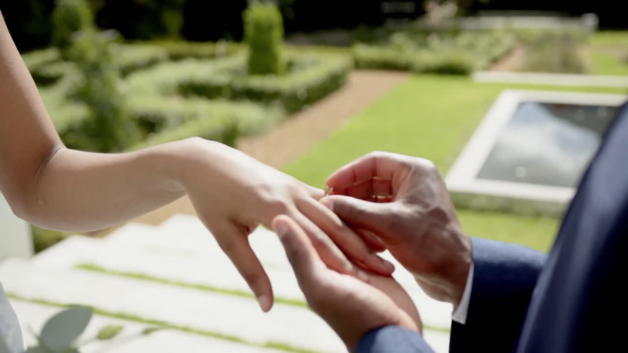 Hands of african american groom putting ring on bride at their wedding in sunny garden, slow motion