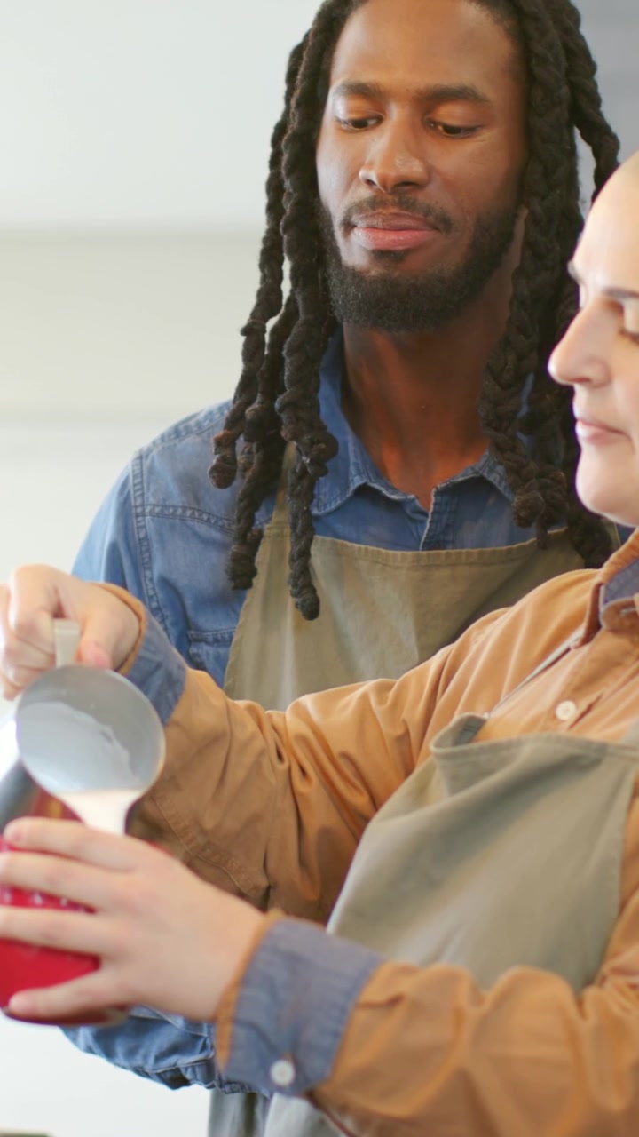 Vertical video: Lifting metal pitcher barista in apron coaching guiding milk pour at cafe, red mug