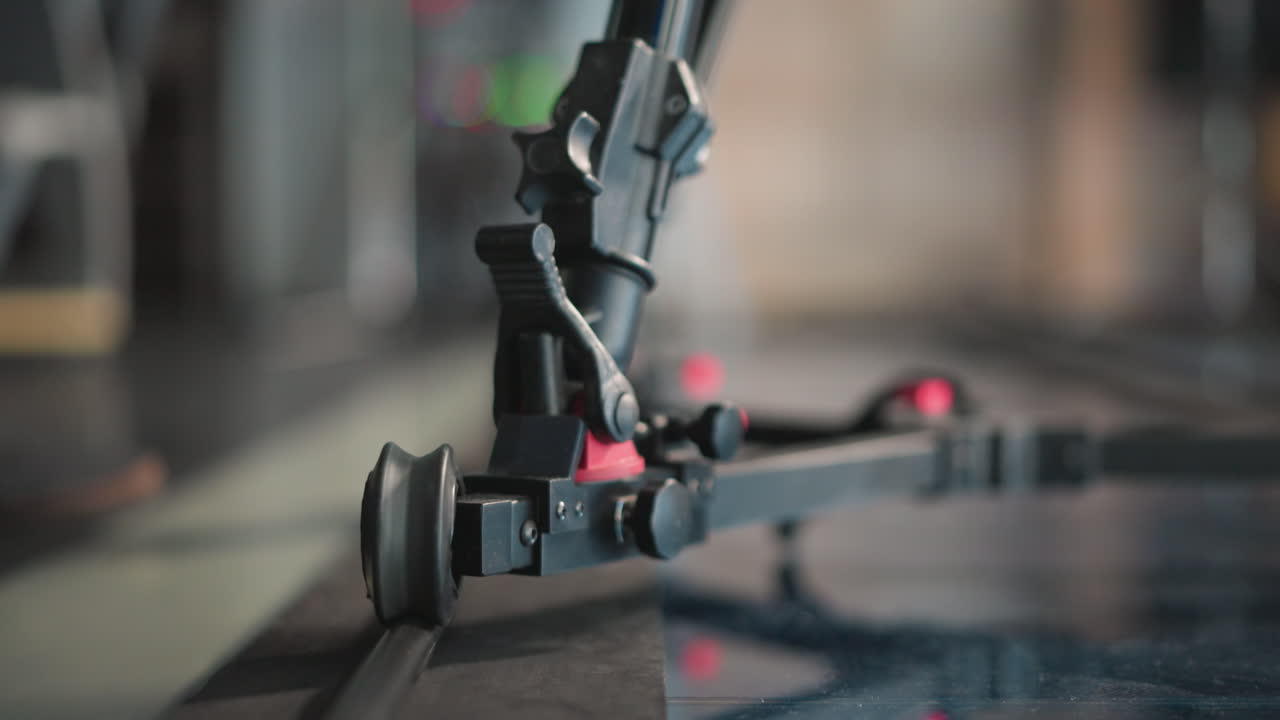 Close-up shot of camera dolly system on track, focusing on wheel and equipment. Reflective studio floor enhances precision of camera rig in action. Red adjustment knob visible