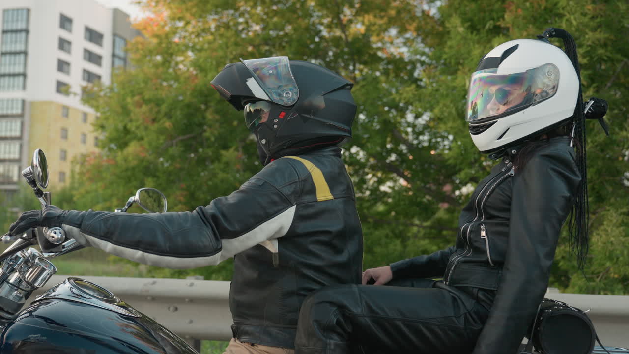 Motorcycle rider carrying passenger adjusts helmet while matching gear on roadside, both wearing black leather jackets with colorful visors, urban buildings and green trees visible in background