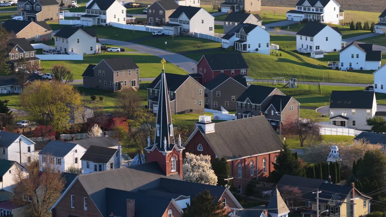 Luftoptagelse i kredsløb af en charmerende amerikansk småby om foråret, med historisk kirketårn omgivet af fredelige kvarterer, blomstrende træer og varmt sollys i rolige boligomgivelser