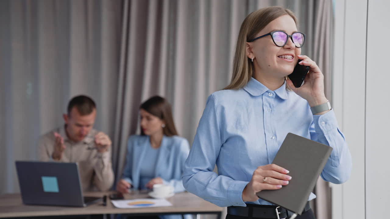 Phone conversation in the office near window. Lady in glasses speaking on the phone holding paper notebook in her hand. Colleagues at desk at backdrop.
