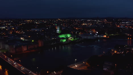 Fly-above-night-city,-green-illuminated-large-building-on-Shannon-river-waterfront.-Limerick,-Ireland