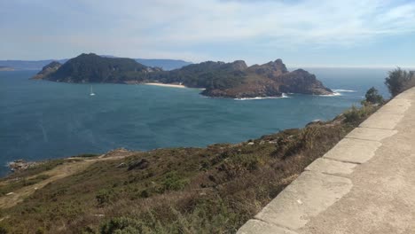 mountainous island with beach and sailboat sailing in front on a sunny day, shot traveling forward, cíes island, pontevedra, galicia, spain