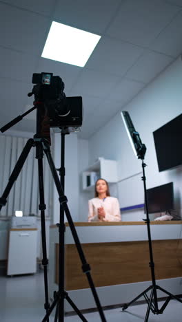 lady with long hair sits at desk giving a speech and gesturing. camera on tripod and daylight lamp are used to record blog content. low angle view. vertical video.