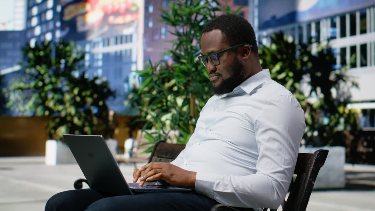 African american male sitting on a bench typing emails on laptop