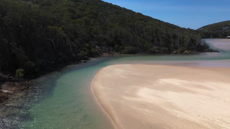 Rising-and-tilting-drone-shot-of-the-Korogoro-Creek-and-coastal-mountains-with-wind-blowing-sand-across-a-sand-bar-at-Hat-Head-New-South-Wales,-Australia