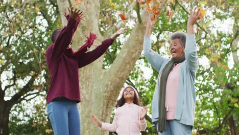 Video-of-happy-african-american-mother,-daughter-and-grandmother-throwing-autumn-leaves-in-garden