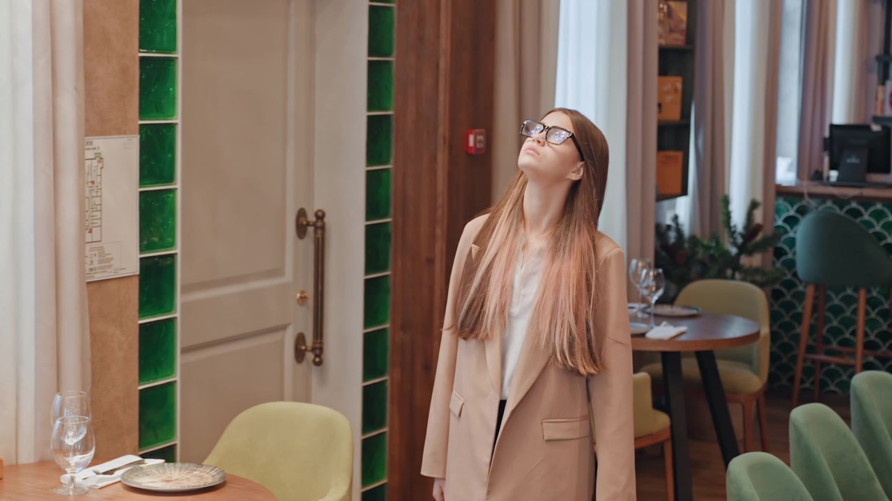 Mujer caucásica inspeccionando los detalles del interior de un restaurante, vistiendo gafas y un blazer beige, con expresión concentrada, evaluando la iluminación y la decoración, con un fondo de barra de azulejos verdes, sillas y mesas puestas, y una ventana.