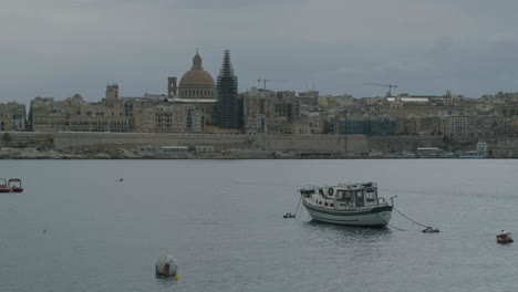 Escena-Pacífica-De-Un-Barco-Aislado-Amarrado-En-El-Puerto-De-Valetta-Lanzando-A-Lo-Largo-De-Las-Olas-Con-La-Ciudad-Vieja-En-El-Fondo,-Malta