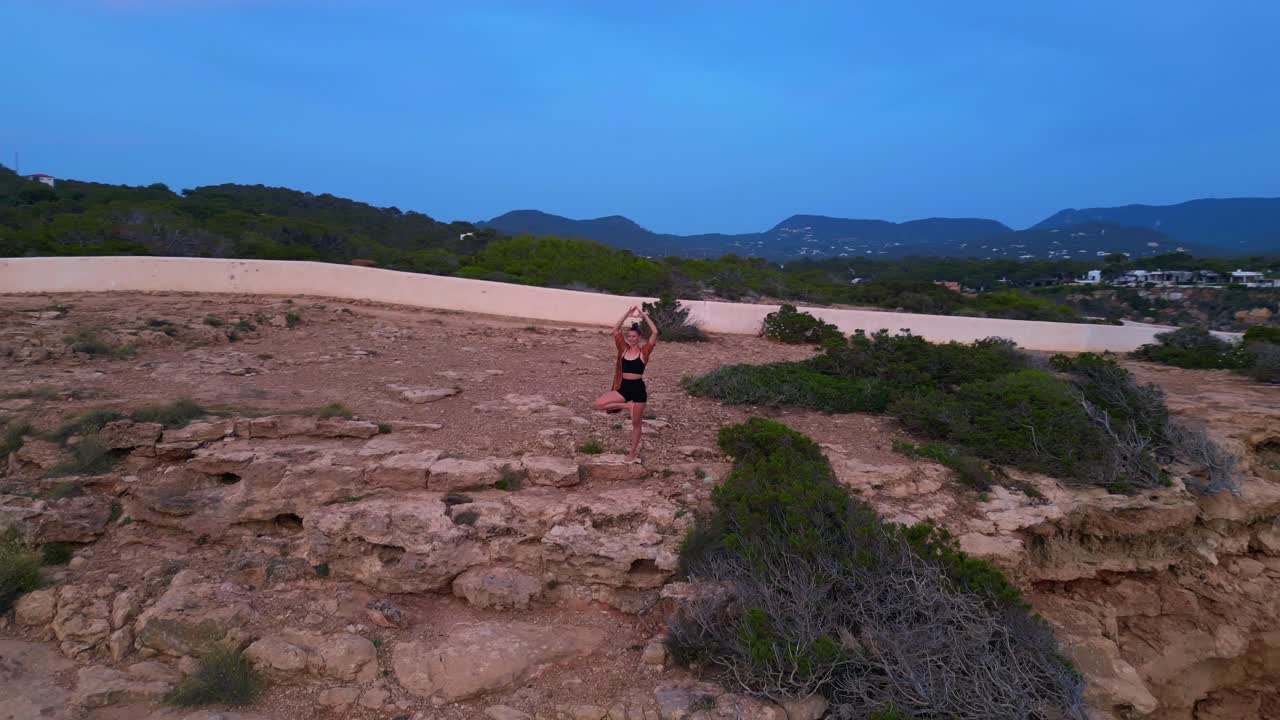 Woman performing a yoga tree pose on a rocky cliff during a serene sunset in Cala Llentia Ibiza. Fantastic aerial view flight circle drone footage