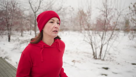 tired adult woman wearing red hoodie and beanie jogging along paved snow-covered path outdoors during winter, surrounded by barren trees and serene park scenery, showing resilience in cold conditions