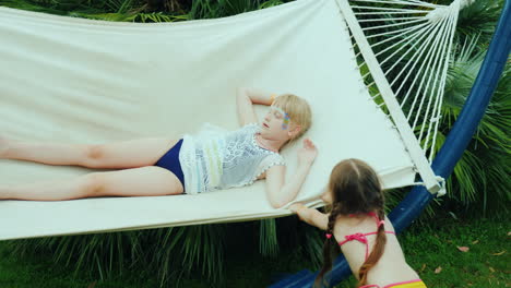 Funny-Children's-Vacation-On-The-Seashore-Two-Sister-Girls-Play-Fun-On-A-Hammock-In-Bathing-Suits