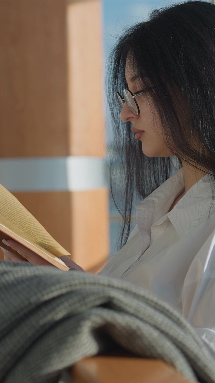 Woman wearing glasses and white shirt. Soft lighting illuminates face and hands holding book gently. Natural daylight creates soft shadows around woman reading outdoors