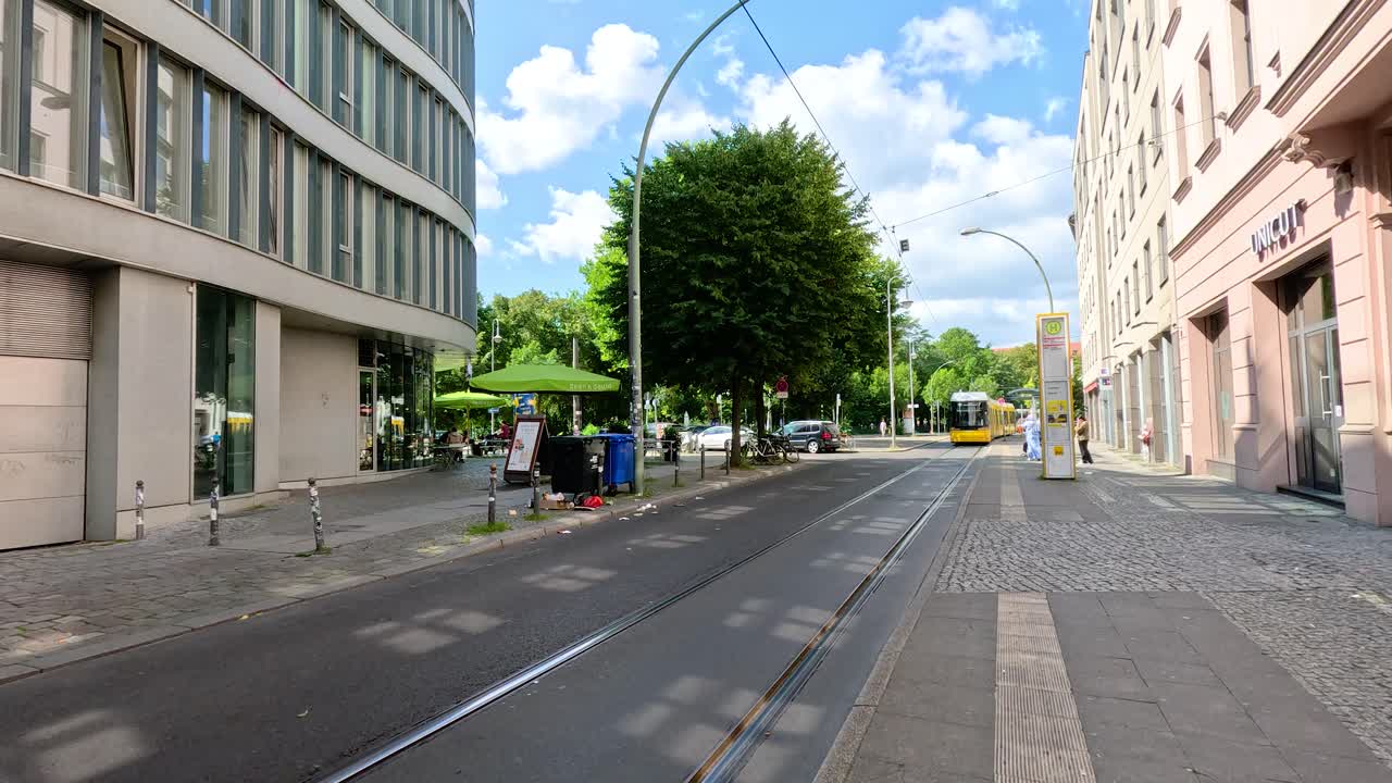 A yellow tram travels down city tracks toward an intersection, passing modern buildings and trees under bright daylight with a clear blue sky