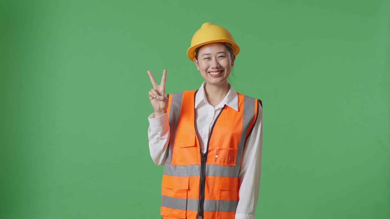 Asian Female Engineer With Safety Helmet Smiling And Showing Peace Gesture While Standing In The Green Screen Background Studio