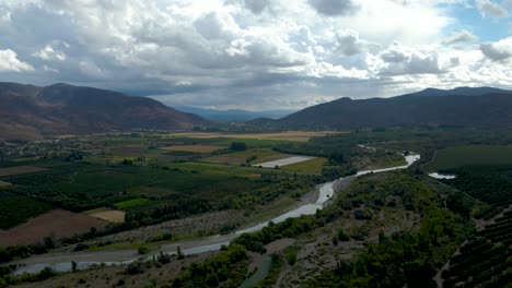 Aerial-orbit-of-green-farm-fields-and-stream-river,-mountains-in-background-on-a-cloudy-day,-Cachapoal-Valley,-south-of-Santiago,-Chile