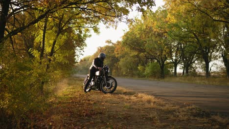 Unrecognizable-stylish-man-in-helmet-and-leather-jacket-coming-up-to-his-bike-and-starting-the-engine-while-standing-on-the-roadside-in-a-sunny-day-in-autumn