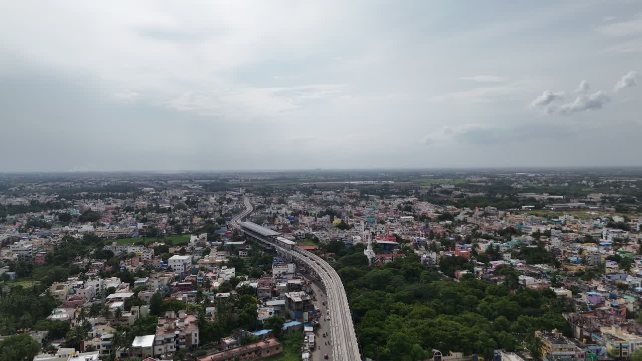 Aerial footage of Iyyappanthangal cityscape with a massive elevated Metro running over the dense, colorful residential sprawl. The shot contrasts old homes with new infrastructure development