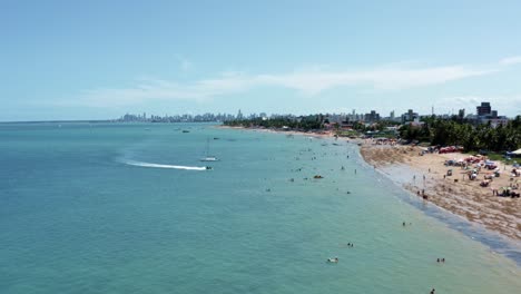 dolly in aerial shot of bessa beach during low tide in the tropical capital city of joao pessoa, paraiba, brazil with people enjoying the ocean and a jet ski approaching the shore on a summer day