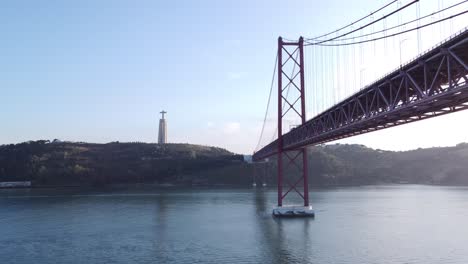 Drone-flying-across-bay-river-in-Portugal-Lisbon-with-famous-bridge-and-monument-of-Jesus-in-Back-Cristo-Rei