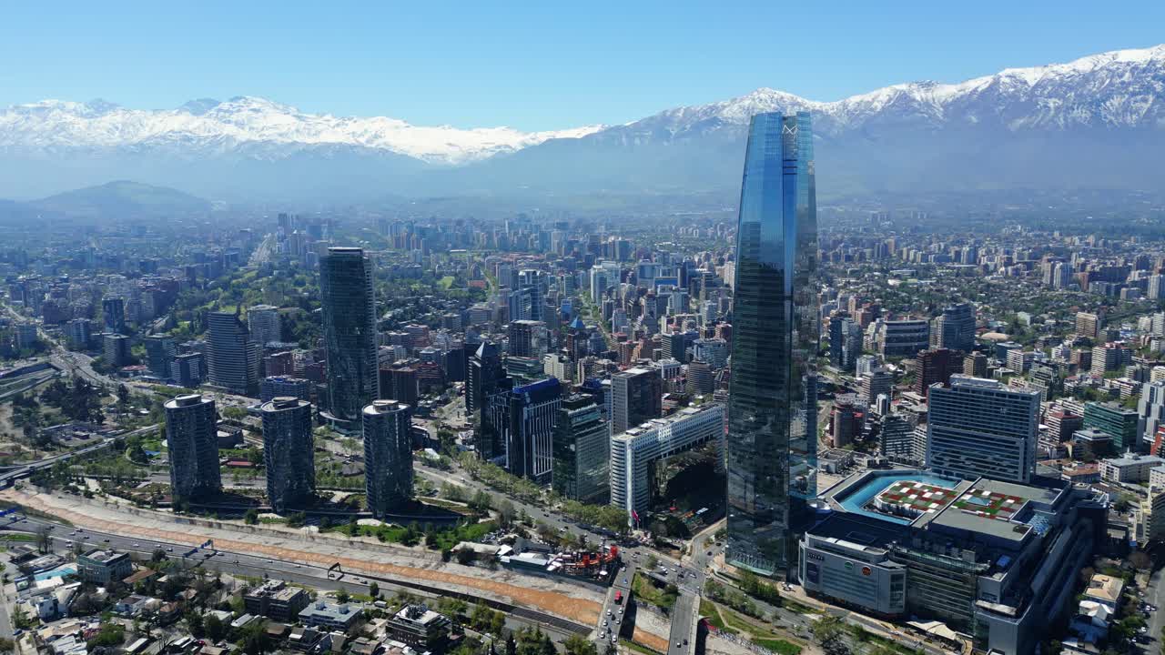 Drone aerial pan and reveal over Costanera skyscrapers and highways, unveiling snowcapped Andes under clear daytime skies. Santiago, Chile