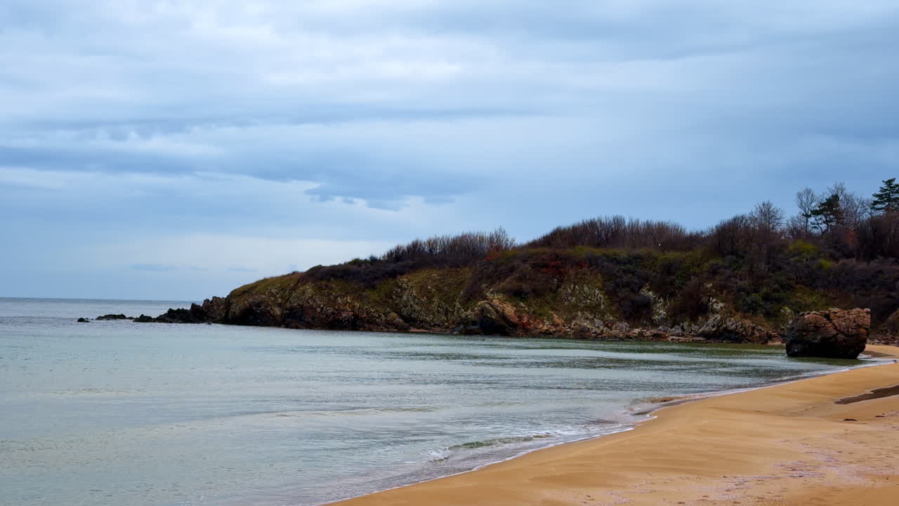 Ocean shore with rocky coast during cloudy weather in early afternoon. Waves touch the sandy beach while a rocky hill stands in the background under gray clouds