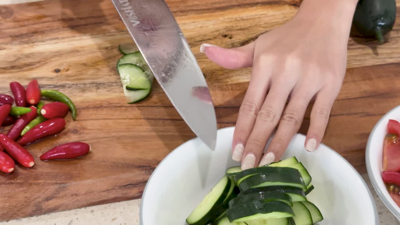 A person slices cucumber and red chili peppers on a wooden cutting board in a bright kitchen, with bowls of fresh vegetables nearby and overhead camera angles