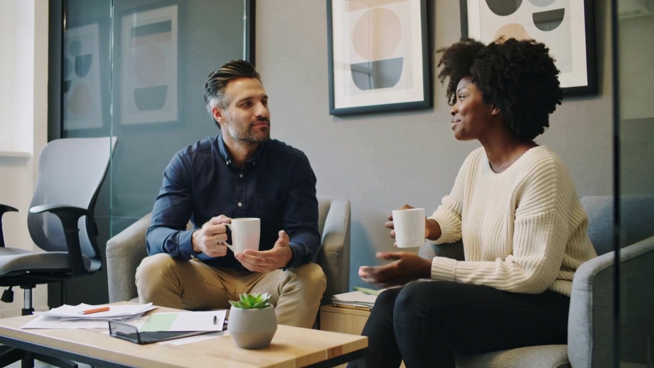 Gesturing male navy shirt starting chat with female cream sweater in office lounge, holding mugs