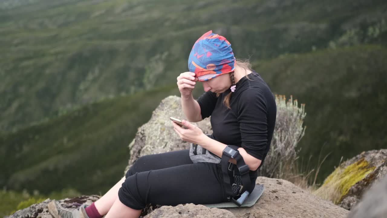 A girl sits on a stone while relaxing on a hike and looks at the phone while adjusting her bandana