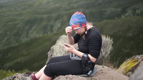 a girl sits on a stone while relaxing on a hike and looks at the phone while adjusting her bandana