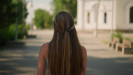 close-up back view of young lady with blonde hair tied in black cyan signet, walking calmly on interlocked pathway featuring scenic background with historical building, benches, lush greenery