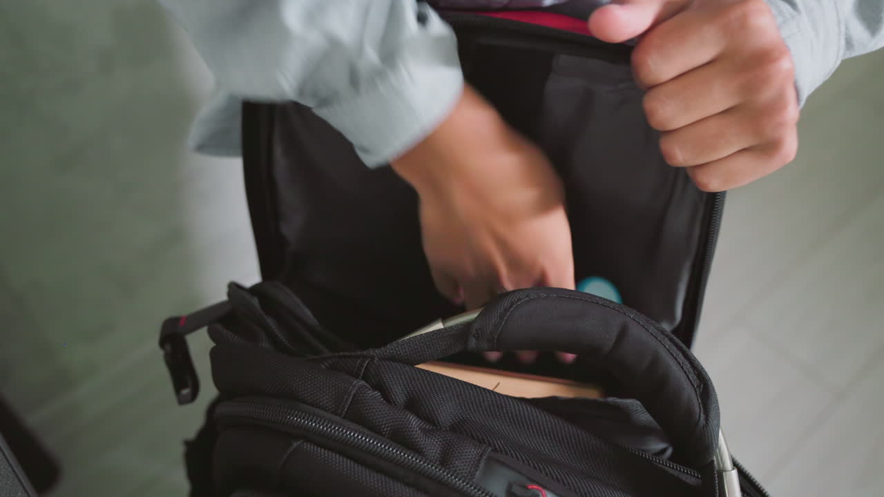 boy in casual shirt holding open black school bag while preparing for school, organizing supplies with focused hands, ready to insert books and materials, indoors near table in clean modern space