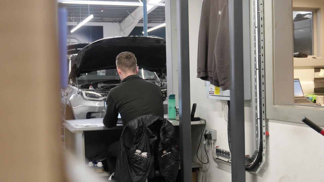 Workshop administrator works at a desk in a busy automotive garage, with a vehicle undergoing maintenance in the background. Scene captures daily operations in a professional repair facility
