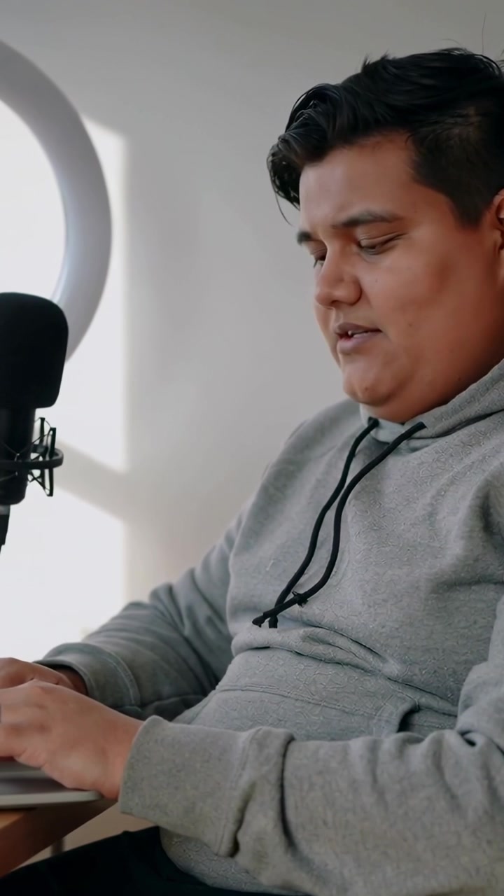 Video verticale: Uomo con felpa con cappuccio che digita e parla al microfono accanto a una ring light, con un laptop, mentre fa podcasting a casa.